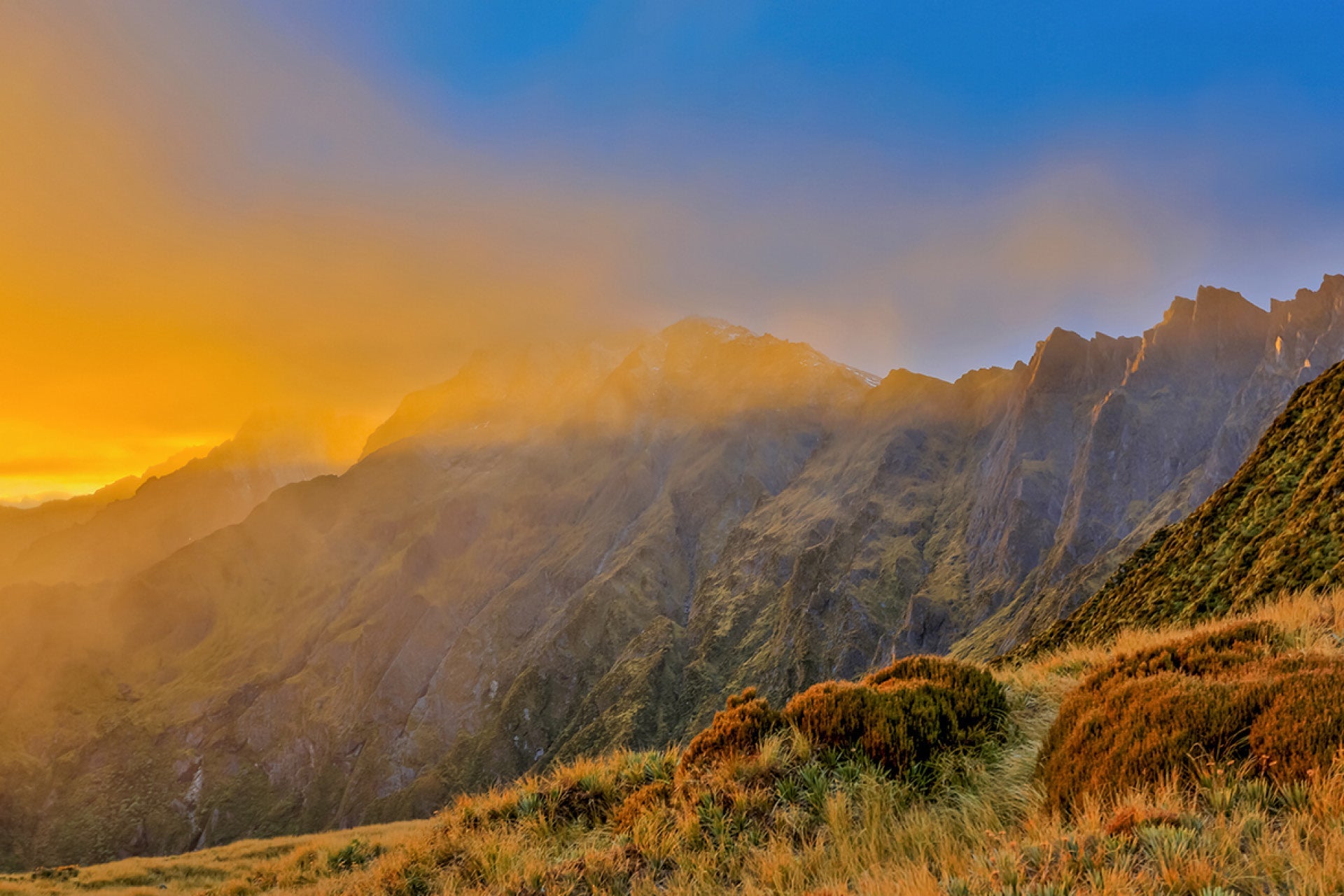Sun igniting misty clouds in an early morning blaze over Fox Glacier on New Zealand's West Coast, leading to Chamois in meadows and glistening Tasman Sea, captured by photographer Steve Couper – perfect for explorers seeking Aotearoa's radiant beauty.