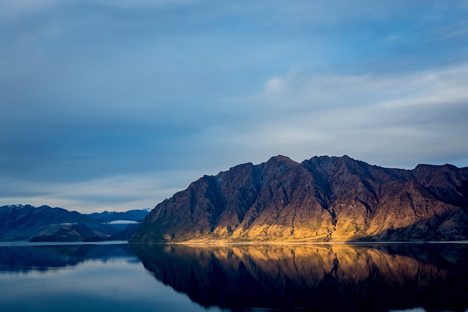 Lake Hawea in Central Otago, New Zealand, where a flat moody afternoon shifts calmly with a shaft of last light illuminating the scene, captured by photographer Steve Couper – ideal for landscape admirers seeking Aotearoa's subtle beauty.